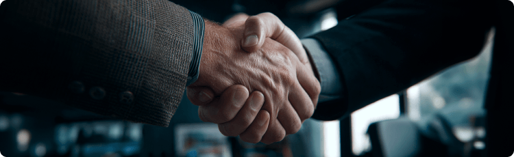 Close-up de um aperto de mÃ£o entre dois homens de fato, com o interior de um escritÃ³rio desfocado ao fundo.