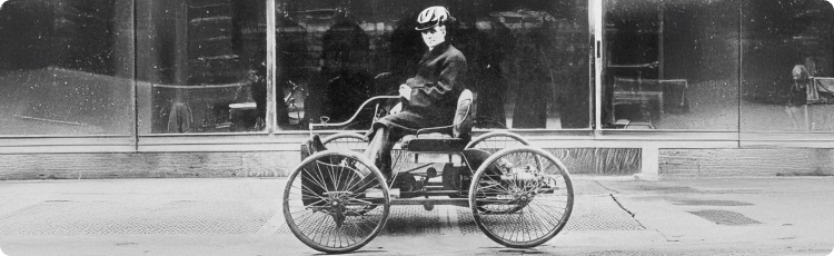 HistorickÃ© trojkolovÃ© motorovÃ© vozidlo riadenÃ© osobou na mestskej ulici, zdÃ´razÅujÃºce ranÃ© technickÃ© inovÃ¡cie relevantnÃ© pre dlhodobÃ½ priemyselnÃ½ a ekonomickÃ½ rozvoj.