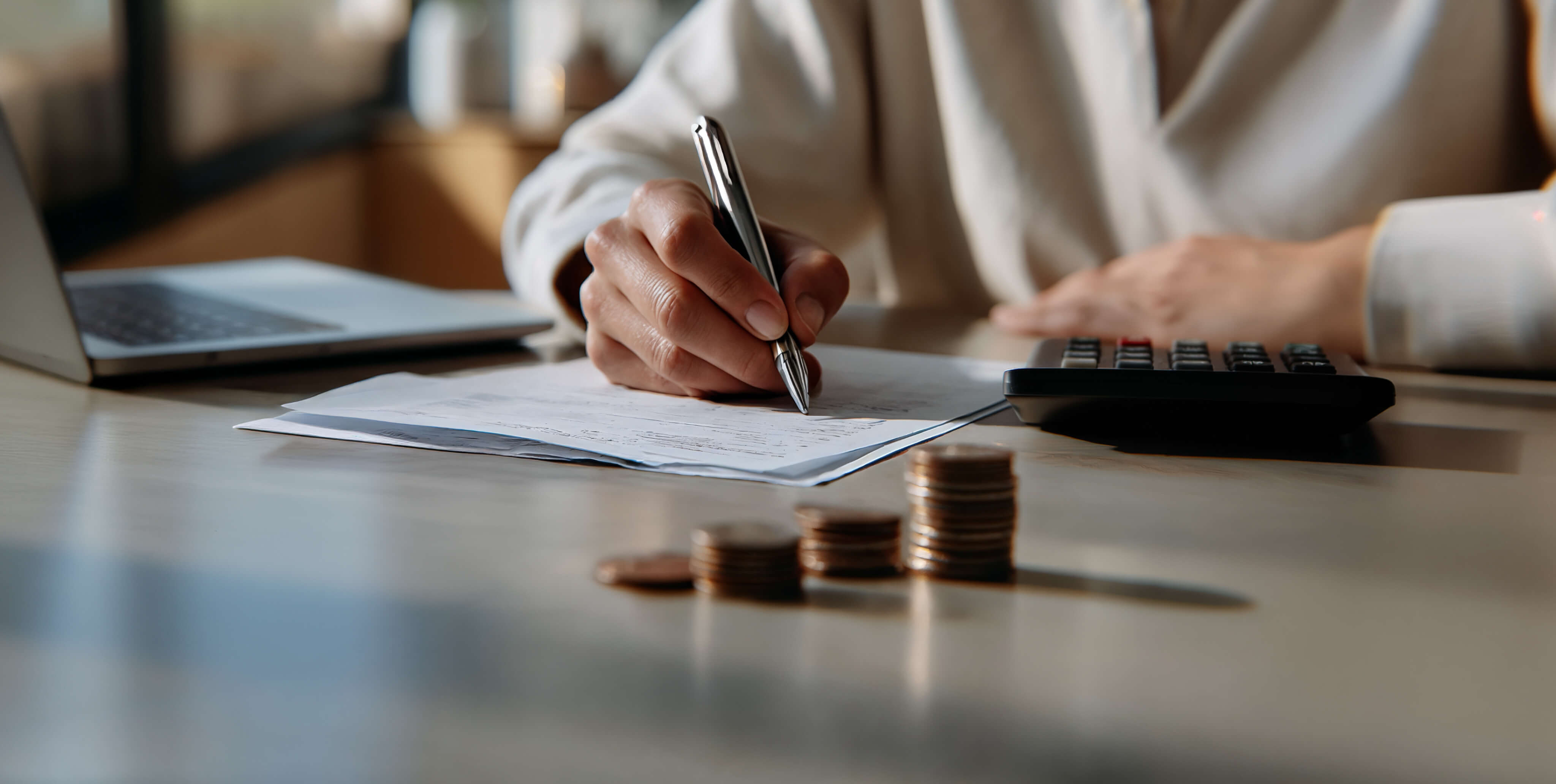 Egy szemÃ©ly pÃ©nzÃ¼gyi dokumentumokra Ã­r egy Ã­rÃ³asztalnÃ¡l, mellette szÃ¡molÃ³gÃ©p, laptop Ã©s egymÃ¡sra rakott Ã©rmÃ©k lÃ¡thatÃ³k, amelyek a szemÃ©lyes kÃ¶ltsÃ©gvetÃ©s tervezÃ©sÃ©t Ã©s a kiadÃ¡skezelÃ©st jelkÃ©pezik.