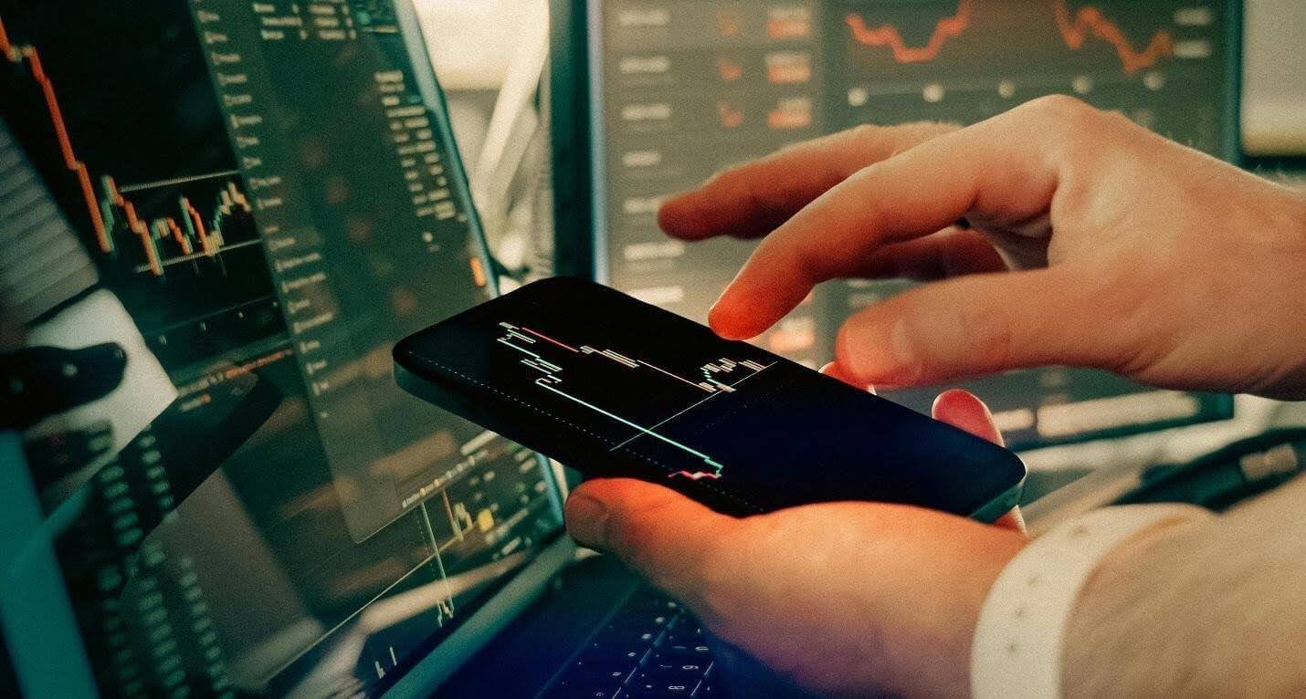Homem a analisar grÃ¡ficos de trading em aplicaÃ§Ã£o mÃ³vel e em desktop