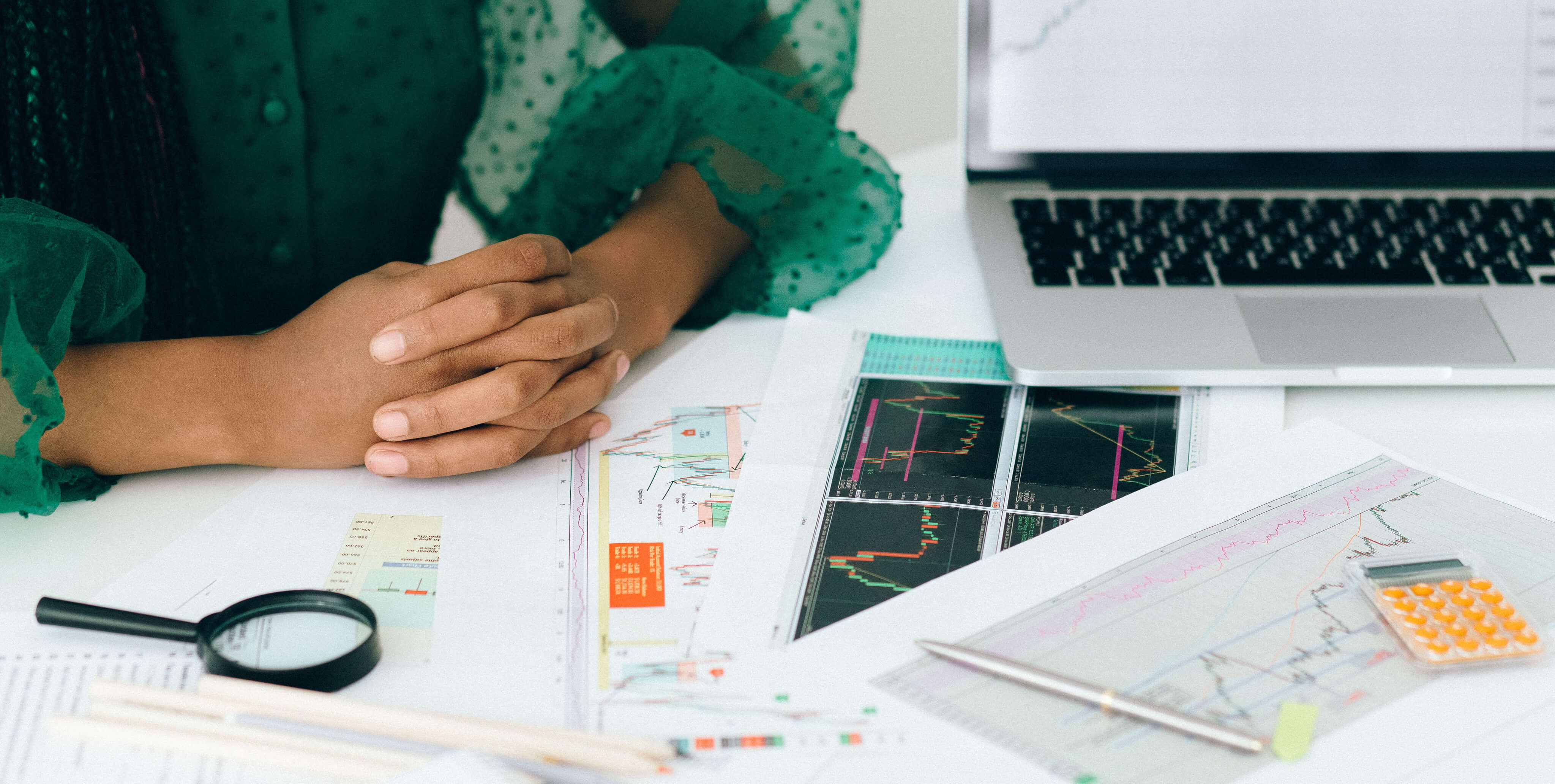 Mulher com as mÃ£os juntas sobre uma mesa com documentos com grÃ¡ficos e um portÃ¡til, ponderando entre investir atravÃ©s de um banco ou de uma corretora