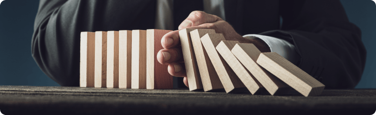 Muž v obleku drží dřevěné bloky jako domino a brání jejich pádu, což symbolizuje řízení rizik v podnikání a investování.