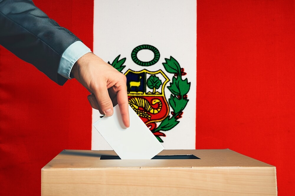 Una mano sostiene una papeleta en una urna con el fondo de la bandera peruana. Las Personas depositan sus votos en una urna con la bandera peruana al fondo. Elecciones en Peru