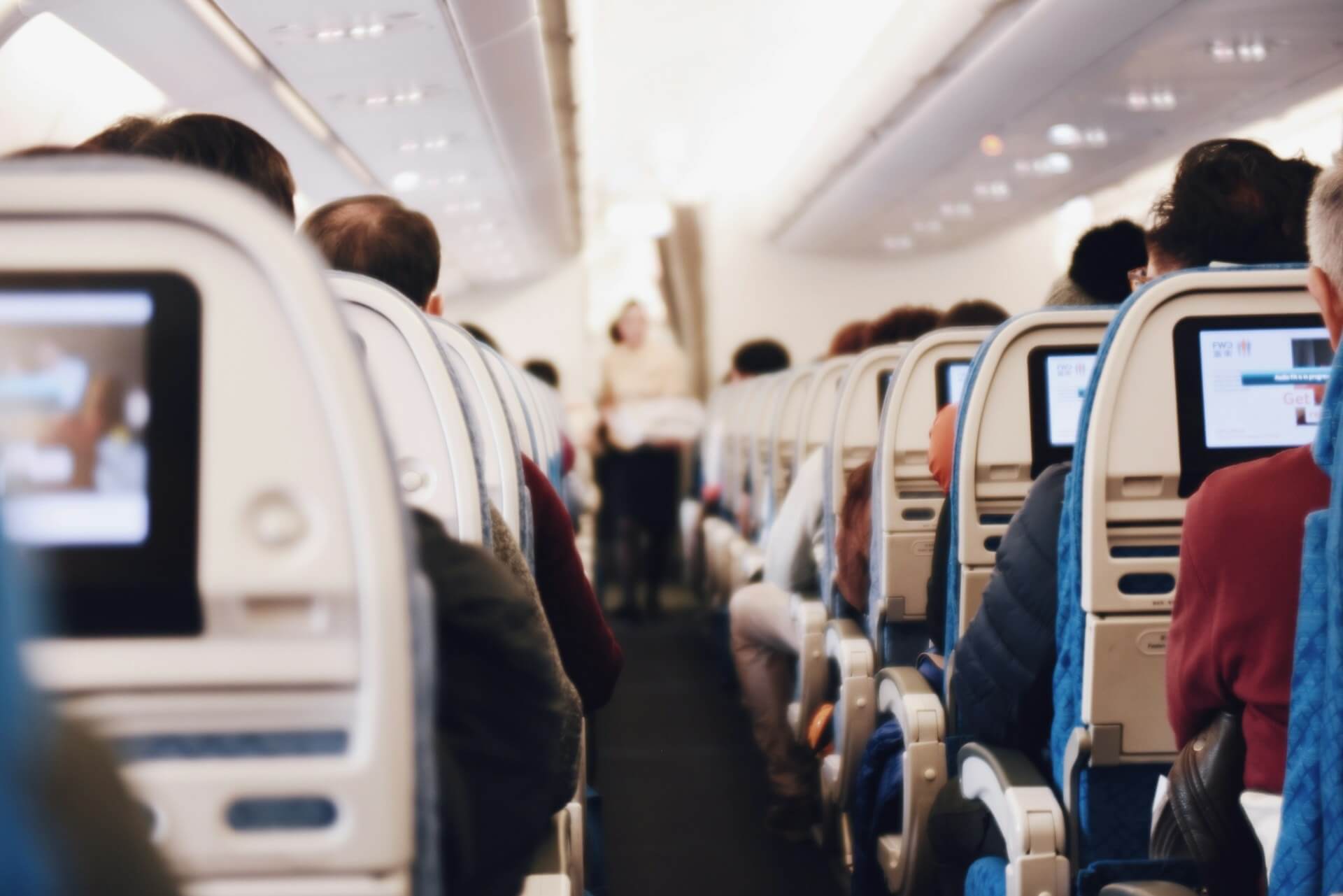 Fotografia de passageiros sentados nos assentos de um aviÃ£o, com uma hospedeira ao fundo desfocada.