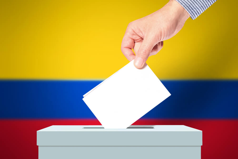 La mano del hombre que pone su voto en las urnas con la bandera de Colombia en segundo plano