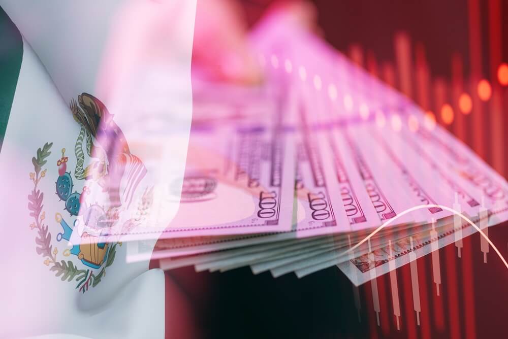 Una mano sostiene una pila de pesos mexicanos y dolares estadounidenses con la bandera mexicana al fondo.